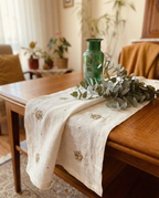 Decorative table setting with a green vase, eucalyptus leaves, and an embroidered table runner on a wooden table. linen table runner