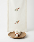 An off-white linen table runner with delicate floral embroidery in muted orange, yellow, and green, styled on a white background with a ceramic plate and napkin.
