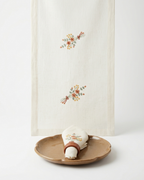 An off-white linen table runner with delicate floral embroidery in muted orange, yellow, and green, styled on a white background with a ceramic plate and napkin.